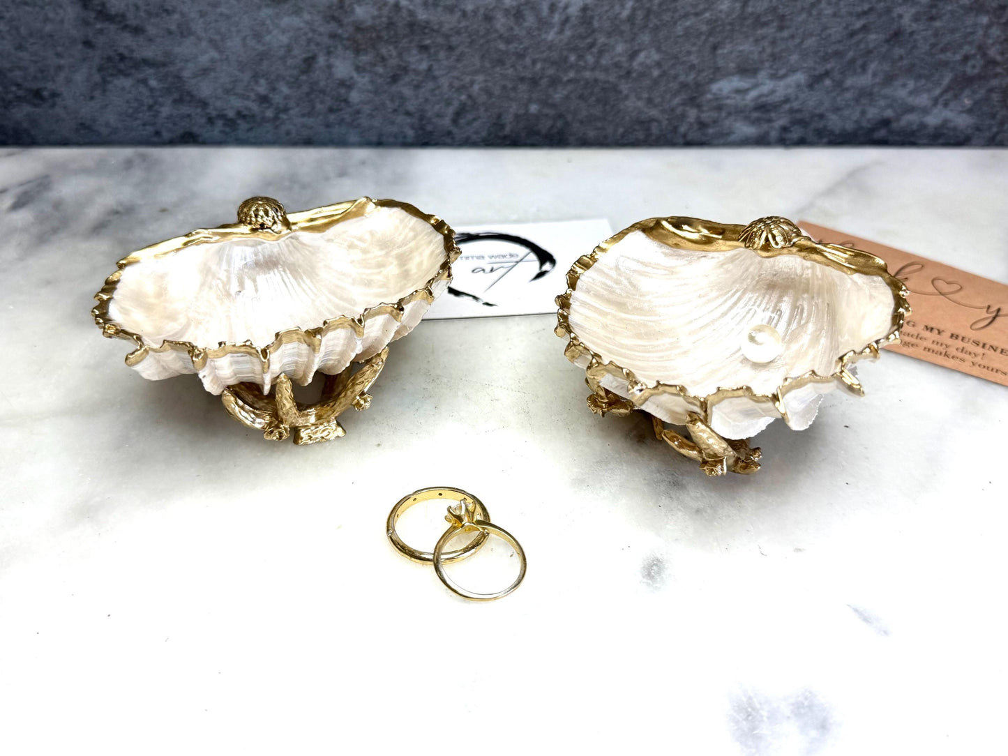 Luxury natural Spiny Cockle shell proposal and wedding ring dish on an ornate gold metal stand, available with or without an inset Swarovski pearl ring holder. Pearlescent shell approximately 8–9cm wide with a hand-gilded liquid gold leaf rim, delicate gold embellishments and crystal-clear resin finish. Handmade luxury jewellery holder, engagement ring dish, bridal keepsake and coastal decor by Emma Wade ART.