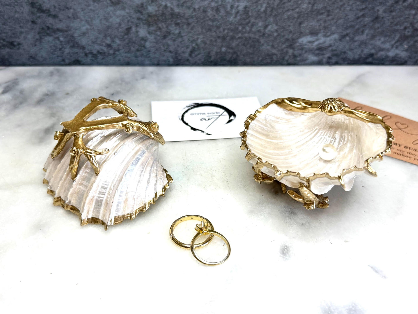 Luxury natural Spiny Cockle shell proposal and wedding ring dish on an ornate gold metal stand, available with or without an inset Swarovski pearl ring holder. Pearlescent shell approximately 8–9cm wide with a hand-gilded liquid gold leaf rim, delicate gold embellishments and crystal-clear resin finish. Handmade luxury jewellery holder, engagement ring dish, bridal keepsake and coastal decor by Emma Wade ART.