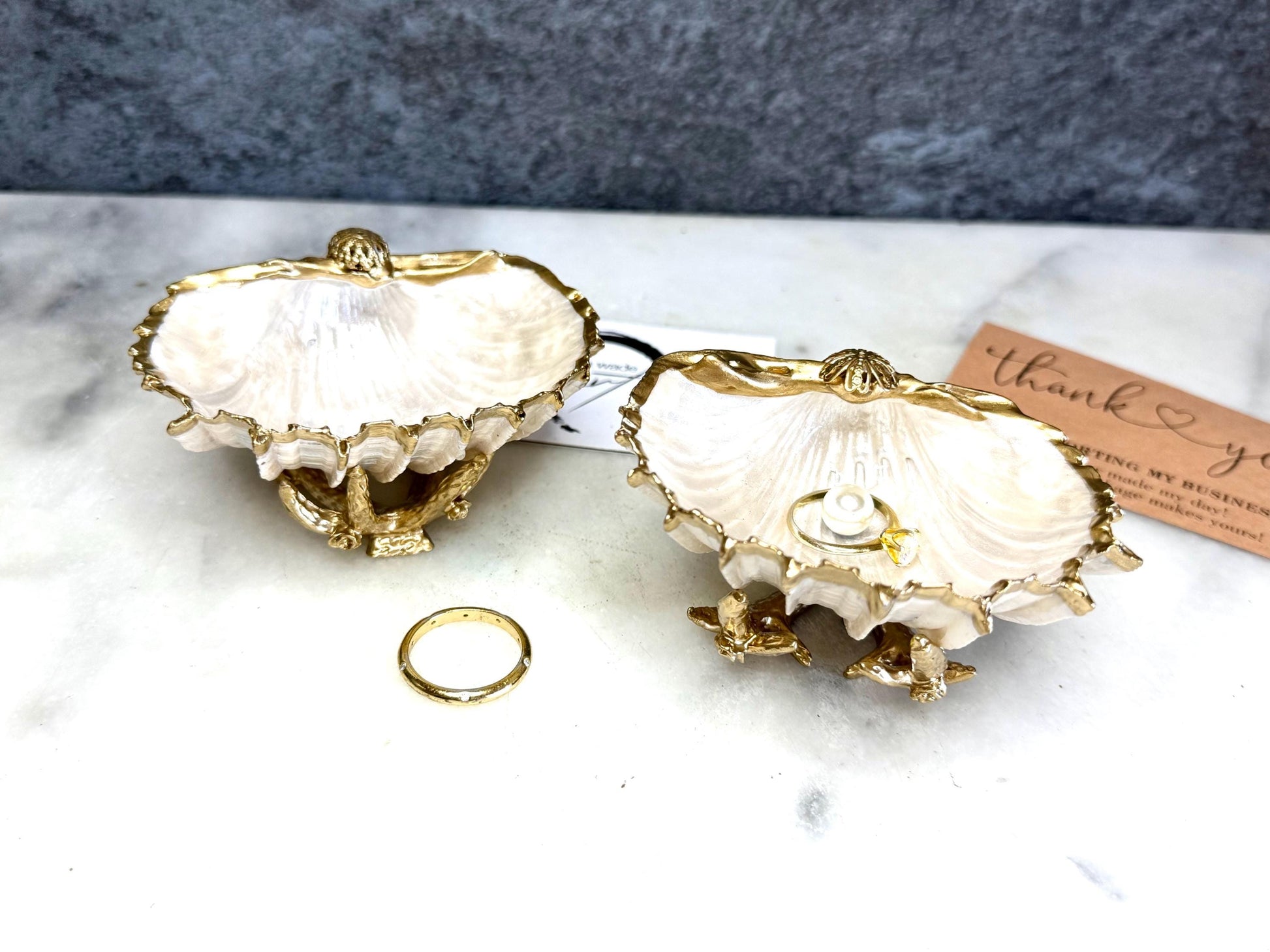 Luxury natural Spiny Cockle shell proposal and wedding ring dish on an ornate gold metal stand, available with or without an inset Swarovski pearl ring holder. Pearlescent shell approximately 8–9cm wide with a hand-gilded liquid gold leaf rim, delicate gold embellishments and crystal-clear resin finish. Handmade luxury jewellery holder, engagement ring dish, bridal keepsake and coastal decor by Emma Wade ART.