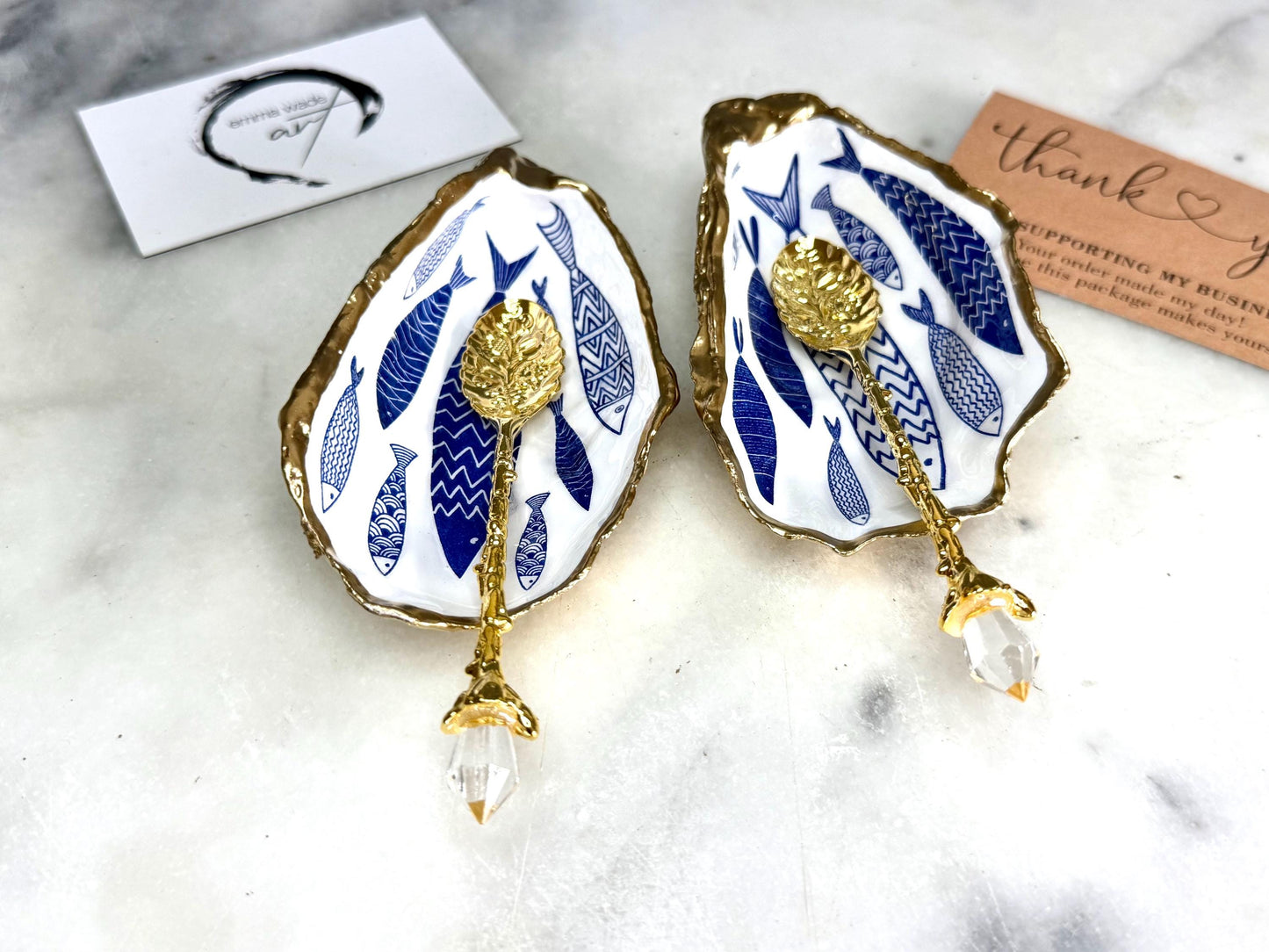 Pair of handmade Dorset oyster shell salt and pepper pinch pots decorated with elegant blue and white fish artwork, finished with hand applied liquid gold leaf edging and sealed in glossy food-safe resin. Coastal shell cruet dishes styled with optional gold crystal spoons on a marble background. Luxury nautical kitchen decor inspired by the sea, perfect for beach house dining, dinner parties, weddings, anniversaries, and coastal home gifts.