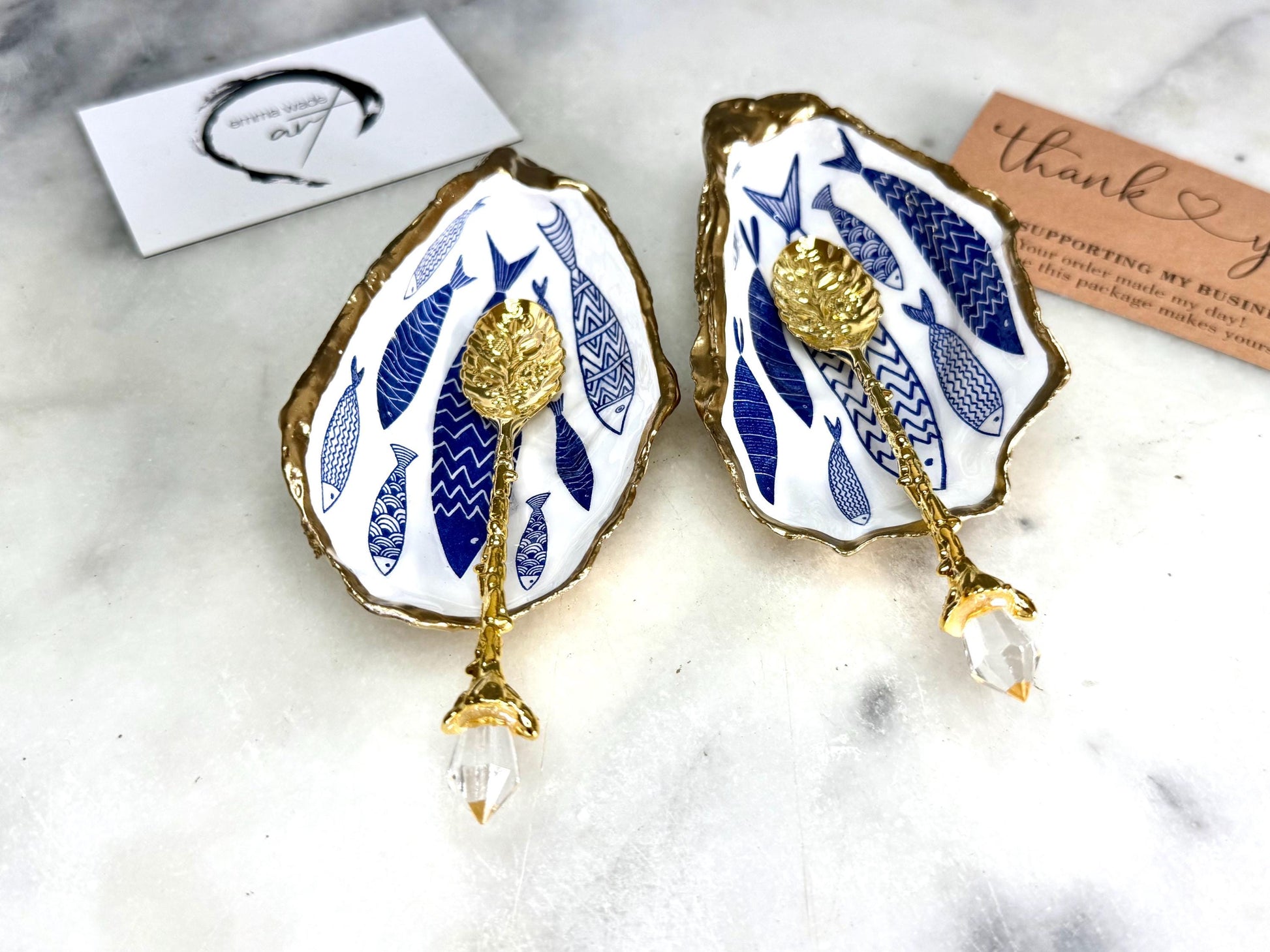 Pair of handmade Dorset oyster shell salt and pepper pinch pots decorated with elegant blue and white fish artwork, finished with hand applied liquid gold leaf edging and sealed in glossy food-safe resin. Coastal shell cruet dishes styled with optional gold crystal spoons on a marble background. Luxury nautical kitchen decor inspired by the sea, perfect for beach house dining, dinner parties, weddings, anniversaries, and coastal home gifts.