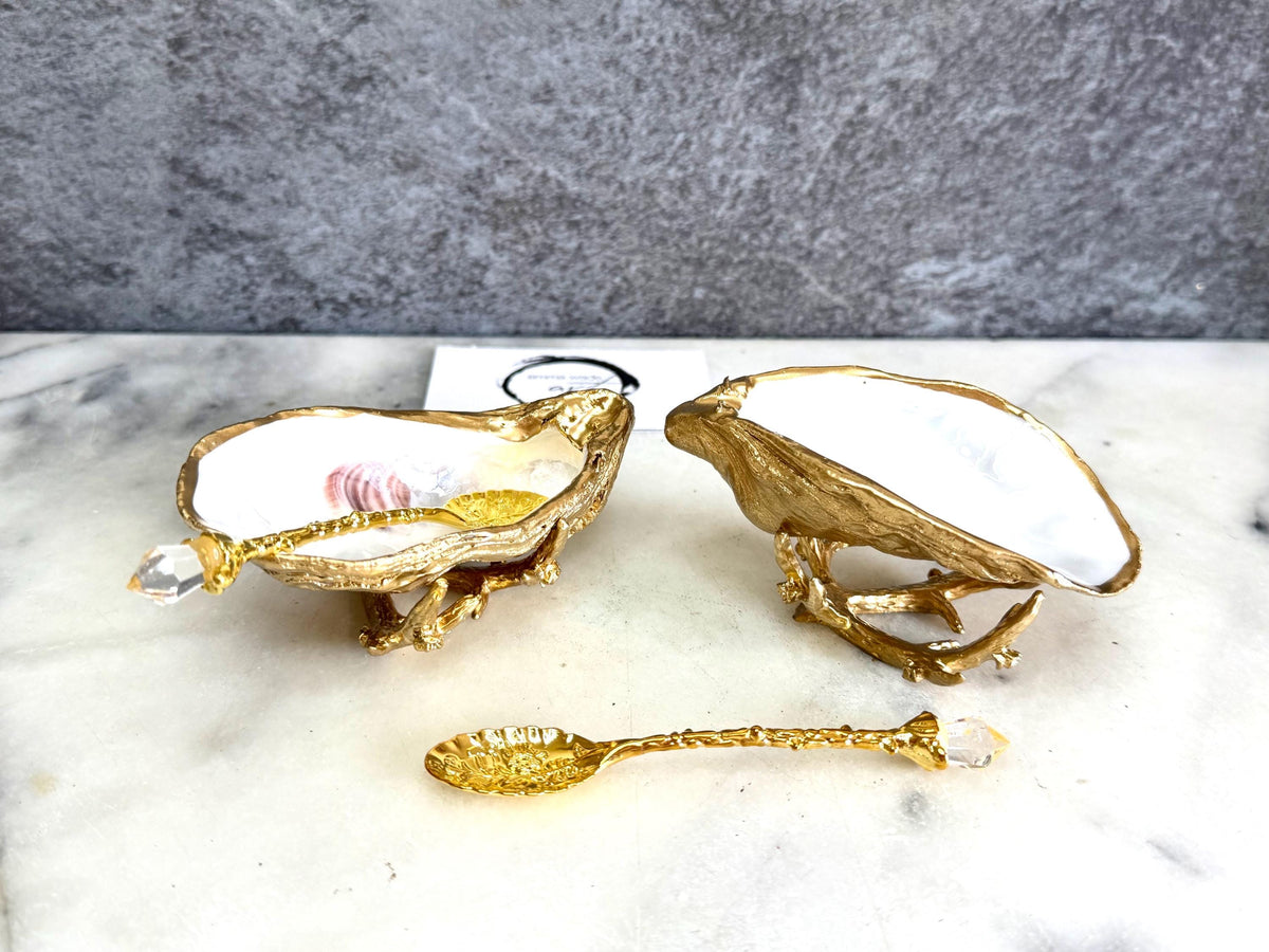 Pair of natural cupped oyster shell salt and pepper dishes displayed on individual driftwood-style gold metal stands. Each oyster shell has a liquid gold leaf rim and glossy resin-sealed pearlescent interior, with a choice of natural gold-highlighted or fully gilded backs. Optional ornate gold spoons with clear crystal ends are shown alongside. Luxury coastal cruet set and wedding or housewarming gift by Emma Wade ART.