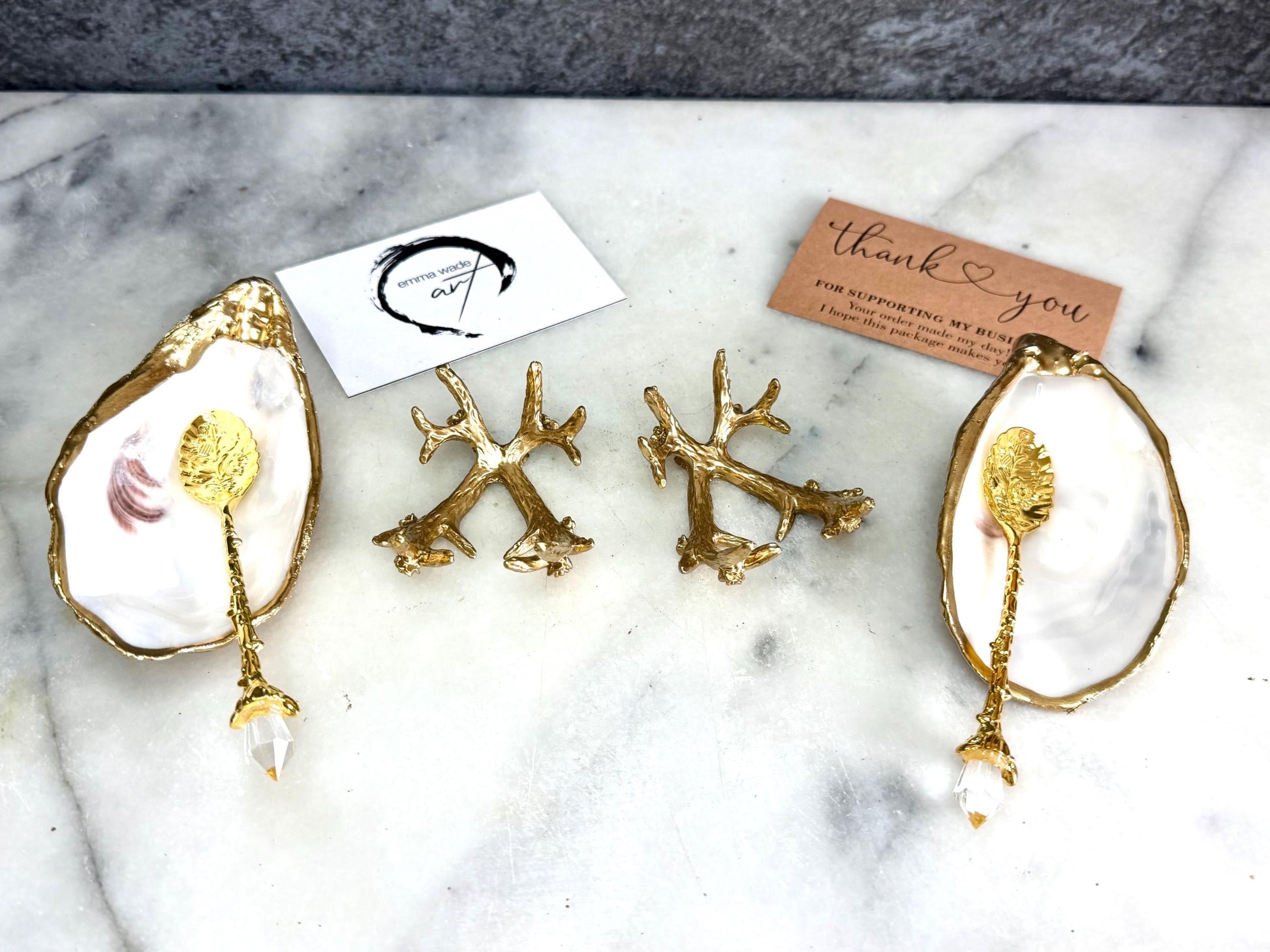 Pair of natural cupped oyster shell salt and pepper dishes displayed on individual driftwood-style gold metal stands. Each oyster shell has a liquid gold leaf rim and glossy resin-sealed pearlescent interior, with a choice of natural gold-highlighted or fully gilded backs. Optional ornate gold spoons with clear crystal ends are shown alongside. Luxury coastal cruet set and wedding or housewarming gift by Emma Wade ART.