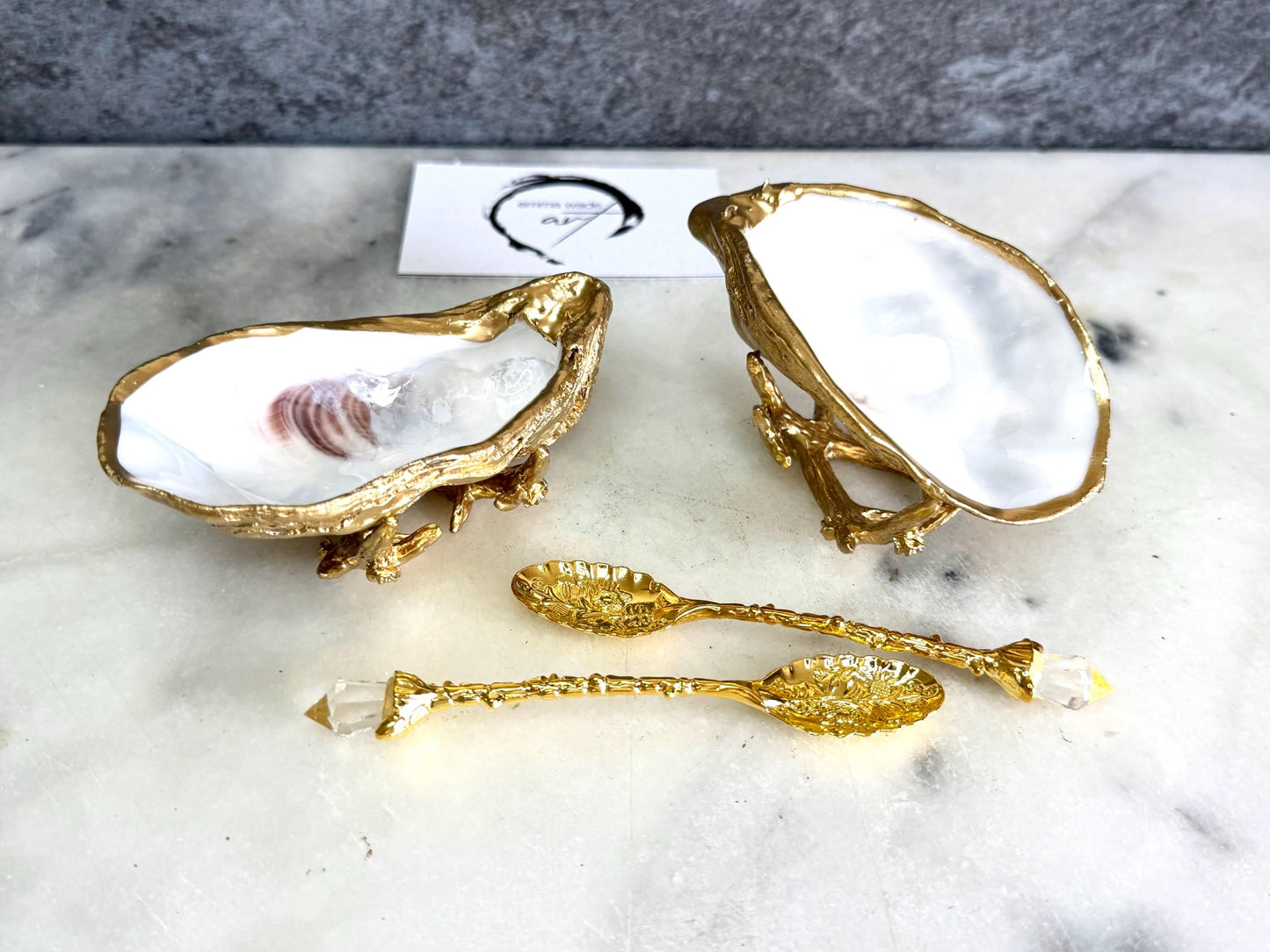 Pair of natural cupped oyster shell salt and pepper dishes displayed on individual driftwood-style gold metal stands. Each oyster shell has a liquid gold leaf rim and glossy resin-sealed pearlescent interior, with a choice of natural gold-highlighted or fully gilded backs. Optional ornate gold spoons with clear crystal ends are shown alongside. Luxury coastal cruet set and wedding or housewarming gift by Emma Wade ART.