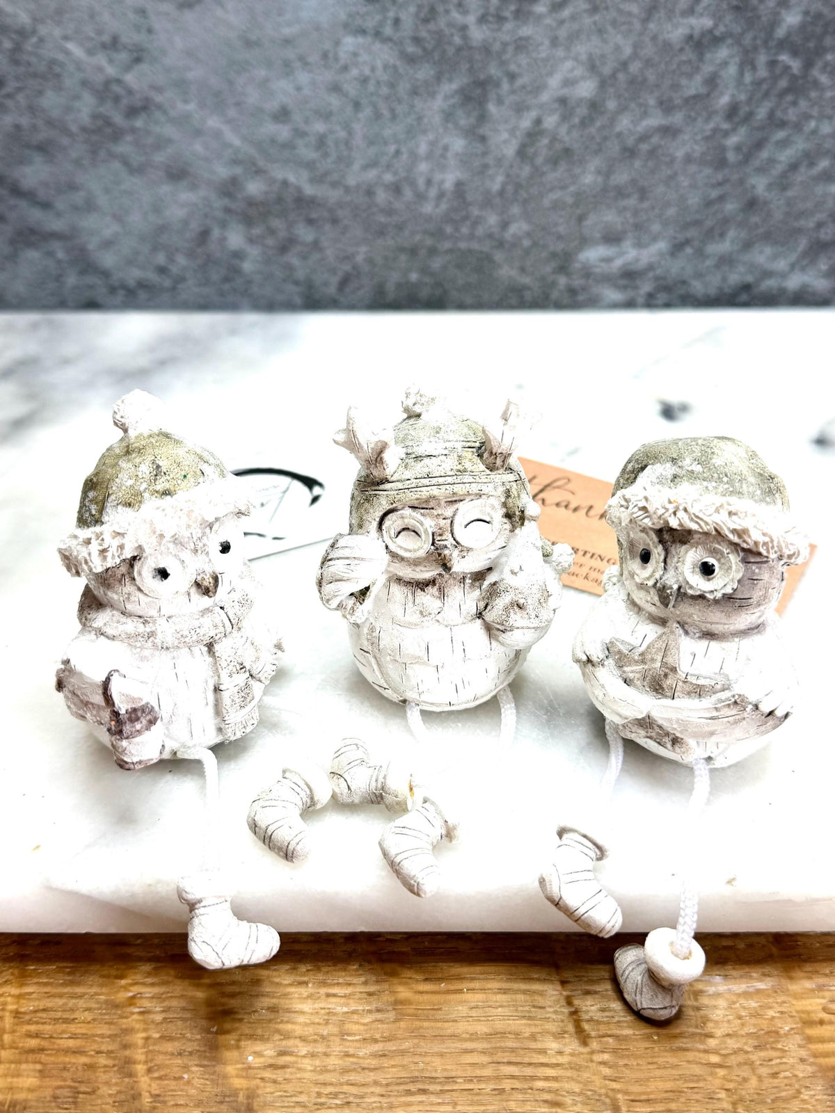 Set of three woodland Christmas owl shelf sitter decorations in soft white, grey and natural tones, featuring winter hats, scarves and festive details. Moulded resin owl ornaments with subtle glitter accents and dangling cord legs with little boots, designed to sit on a shelf, mantelpiece or windowsill. Festive woodland Christmas décor and owl lover gift.