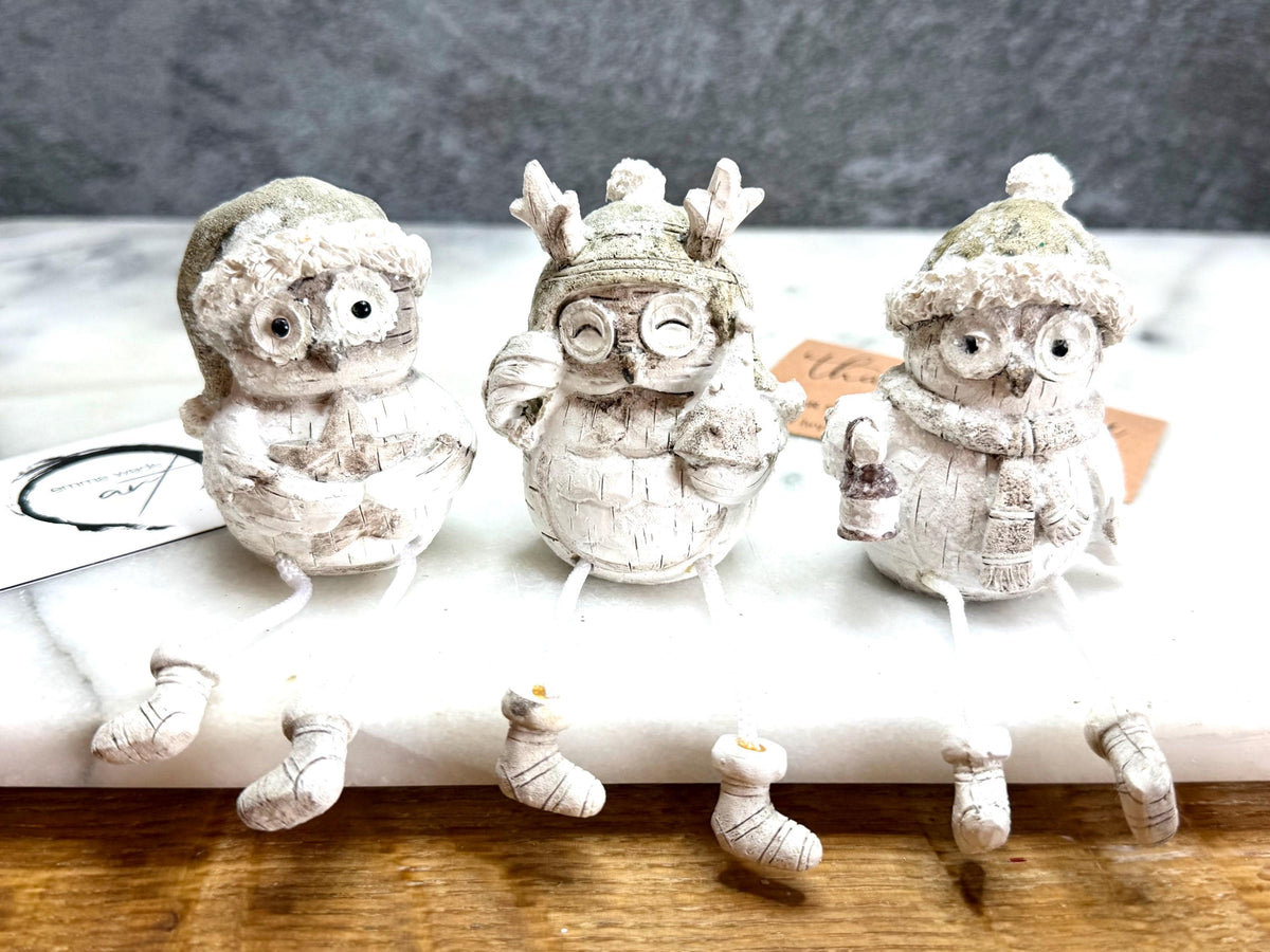 Set of three woodland Christmas owl shelf sitter decorations in soft white, grey and natural tones, featuring winter hats, scarves and festive details. Moulded resin owl ornaments with subtle glitter accents and dangling cord legs with little boots, designed to sit on a shelf, mantelpiece or windowsill. Festive woodland Christmas décor and owl lover gift.