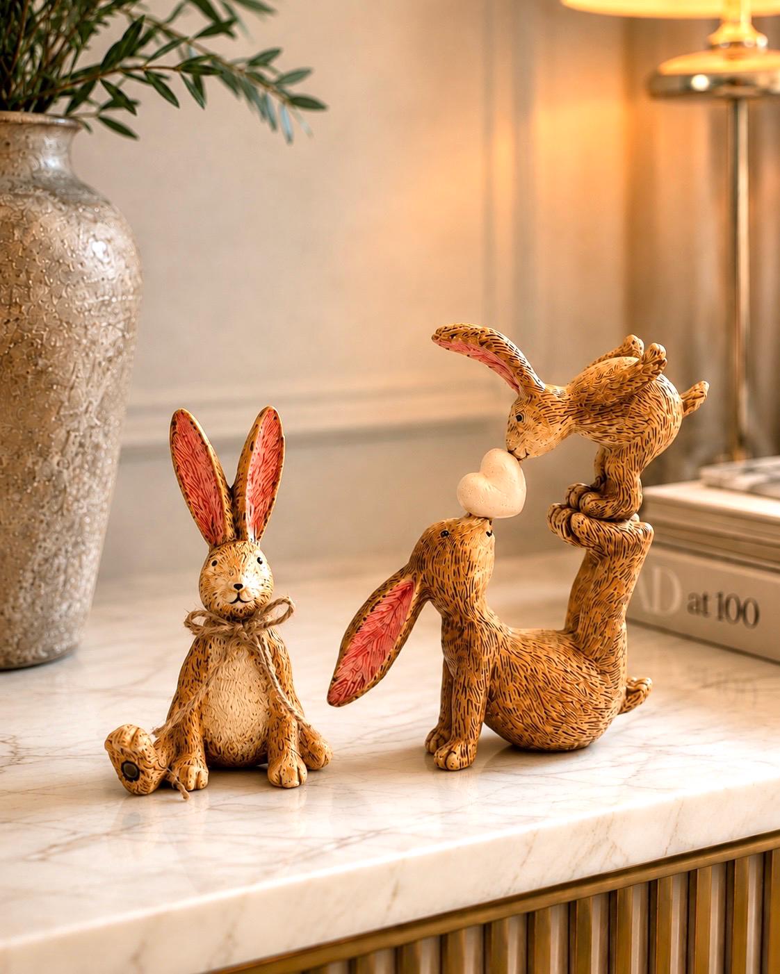 Hand finished polyresin Christmas bunny ornaments with rustic jute bows and soft pink inner ears. Decorative rabbit shelf ornaments shown as a seated bunny and two bunnies kissing, available individually or as a set. Festive woodland-style decor for Christmas shelves, mantelpieces, cottage interiors and rabbit lover gifts.