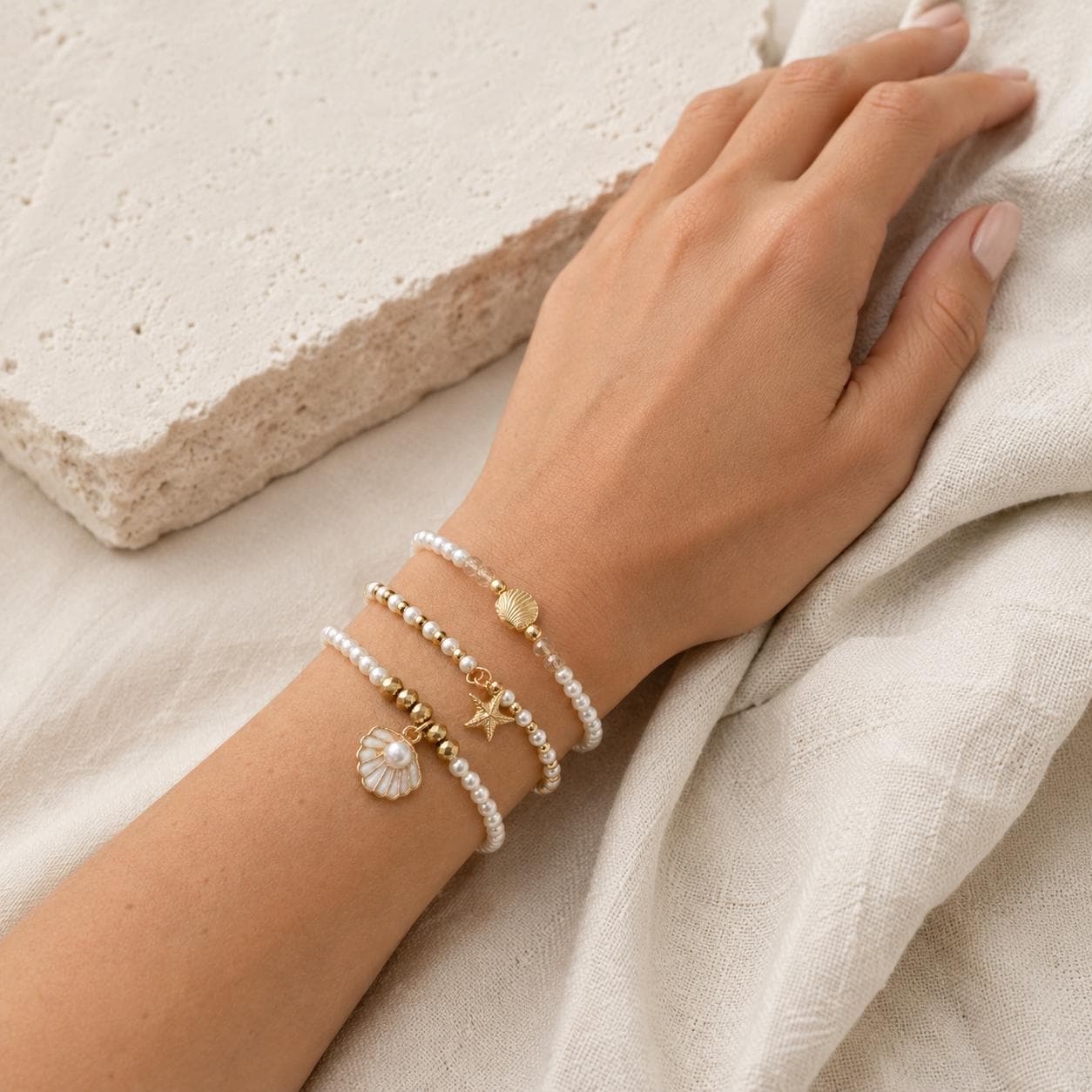 Set of three delicate coastal stretch bracelets featuring small ivory faux pearls and gold-tone seaside details. The coordinating jewellery set includes a blush enamel-style scallop shell charm with pearl centre, a textured gold starfish charm and a ridged gold shell centrepiece with pale champagne faceted beads. Lightweight 16 cm elasticated bohemian costume jewellery for coastal gifts, Christmas presents, stocking fillers, holidays, beach weddings, bridesmaids and gifts for her.