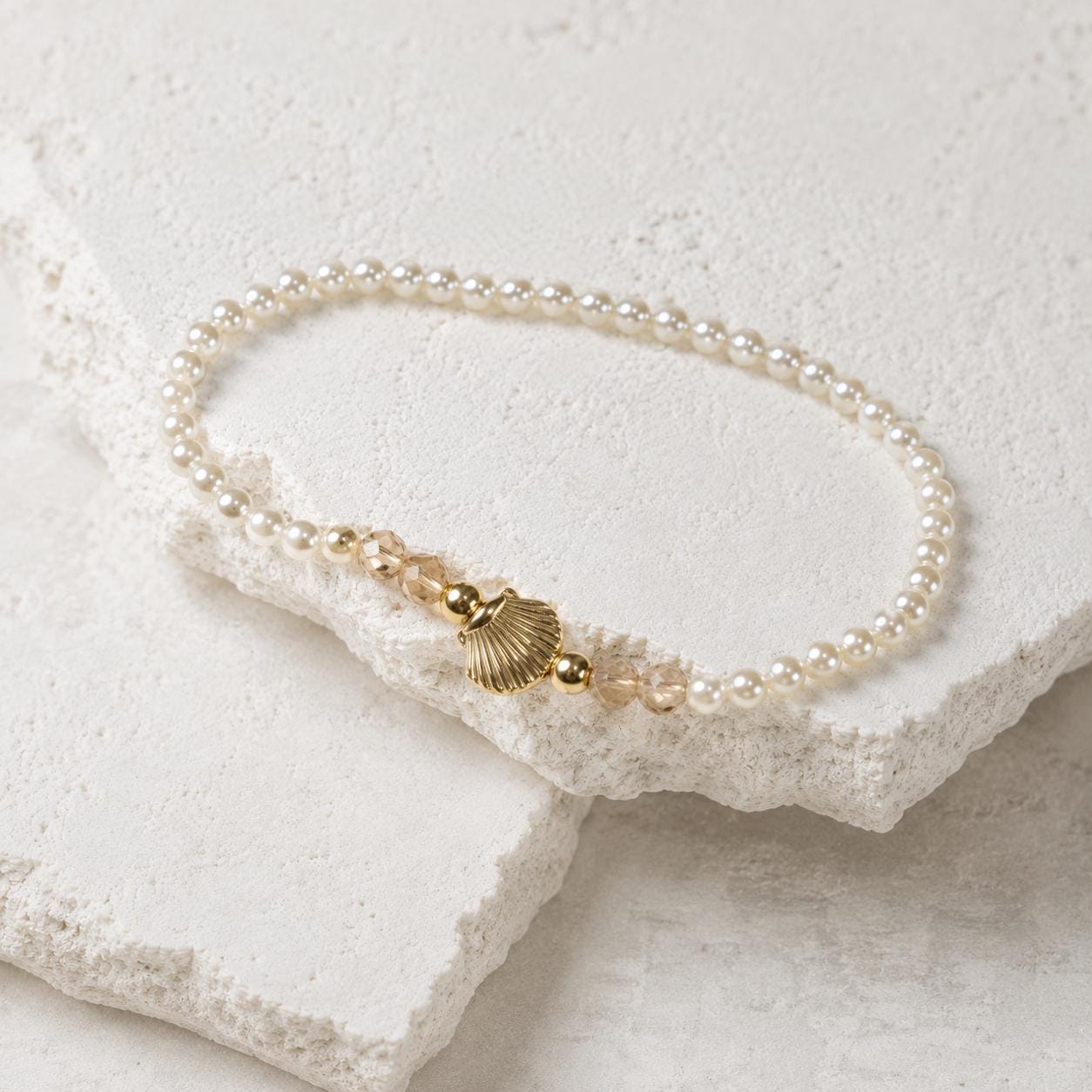 Set of three delicate coastal stretch bracelets featuring small ivory faux pearls and gold-tone seaside details. The coordinating jewellery set includes a blush enamel-style scallop shell charm with pearl centre, a textured gold starfish charm and a ridged gold shell centrepiece with pale champagne faceted beads. Lightweight 16 cm elasticated bohemian costume jewellery for coastal gifts, Christmas presents, stocking fillers, holidays, beach weddings, bridesmaids and gifts for her.