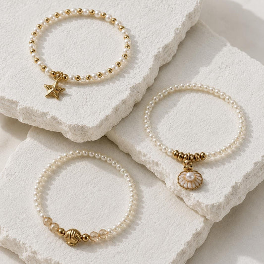 Set of three delicate coastal stretch bracelets featuring small ivory faux pearls and gold-tone seaside details. The coordinating jewellery set includes a blush enamel-style scallop shell charm with pearl centre, a textured gold starfish charm and a ridged gold shell centrepiece with pale champagne faceted beads. Lightweight 16 cm elasticated bohemian costume jewellery for coastal gifts, Christmas presents, stocking fillers, holidays, beach weddings, bridesmaids and gifts for her.
