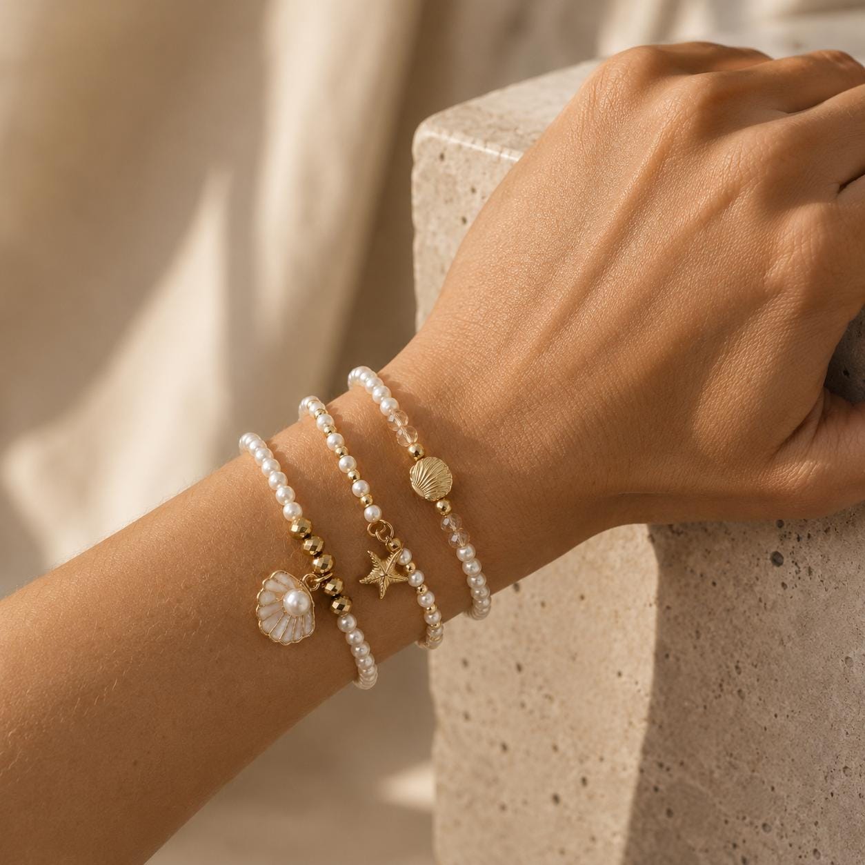 Set of three delicate coastal stretch bracelets featuring small ivory faux pearls and gold-tone seaside details. The coordinating jewellery set includes a blush enamel-style scallop shell charm with pearl centre, a textured gold starfish charm and a ridged gold shell centrepiece with pale champagne faceted beads. Lightweight 16 cm elasticated bohemian costume jewellery for coastal gifts, Christmas presents, stocking fillers, holidays, beach weddings, bridesmaids and gifts for her.