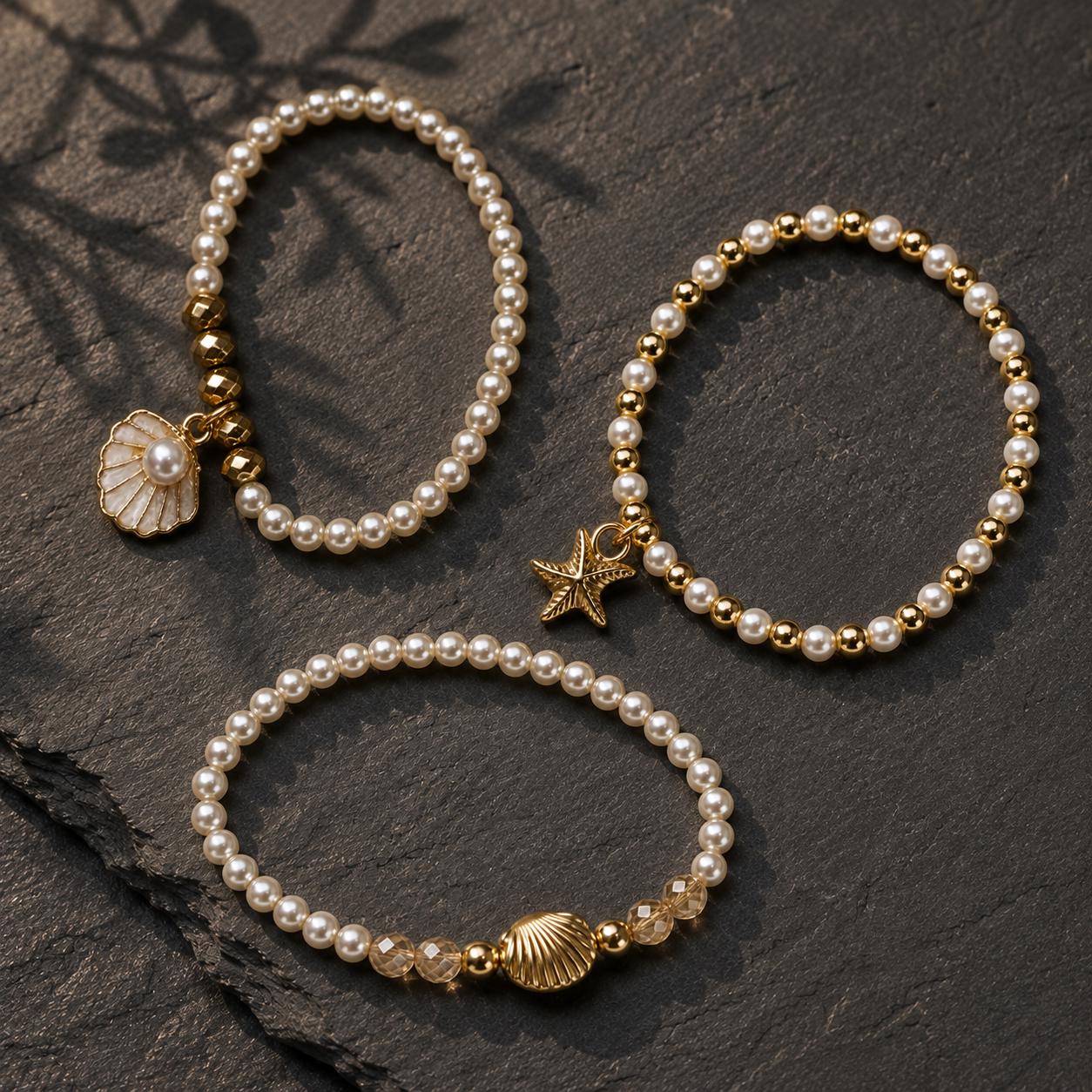 Set of three delicate coastal stretch bracelets featuring small ivory faux pearls and gold-tone seaside details. The coordinating jewellery set includes a blush enamel-style scallop shell charm with pearl centre, a textured gold starfish charm and a ridged gold shell centrepiece with pale champagne faceted beads. Lightweight 16 cm elasticated bohemian costume jewellery for coastal gifts, Christmas presents, stocking fillers, holidays, beach weddings, bridesmaids and gifts for her.