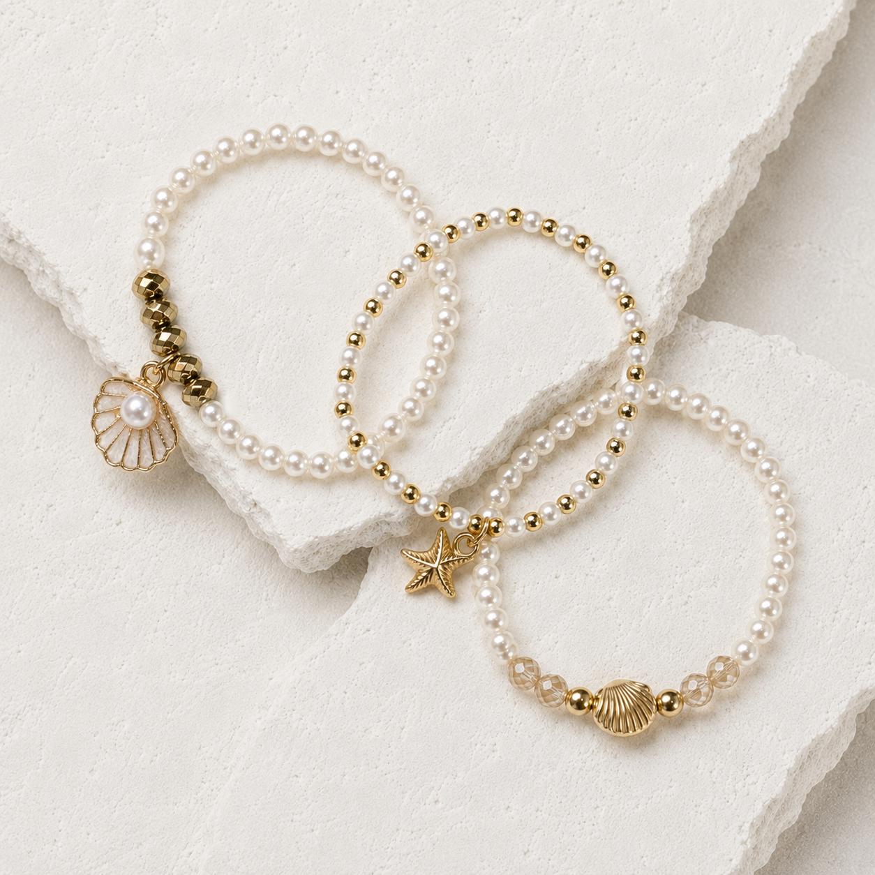 Set of three delicate coastal stretch bracelets featuring small ivory faux pearls and gold-tone seaside details. The coordinating jewellery set includes a blush enamel-style scallop shell charm with pearl centre, a textured gold starfish charm and a ridged gold shell centrepiece with pale champagne faceted beads. Lightweight 16 cm elasticated bohemian costume jewellery for coastal gifts, Christmas presents, stocking fillers, holidays, beach weddings, bridesmaids and gifts for her.
