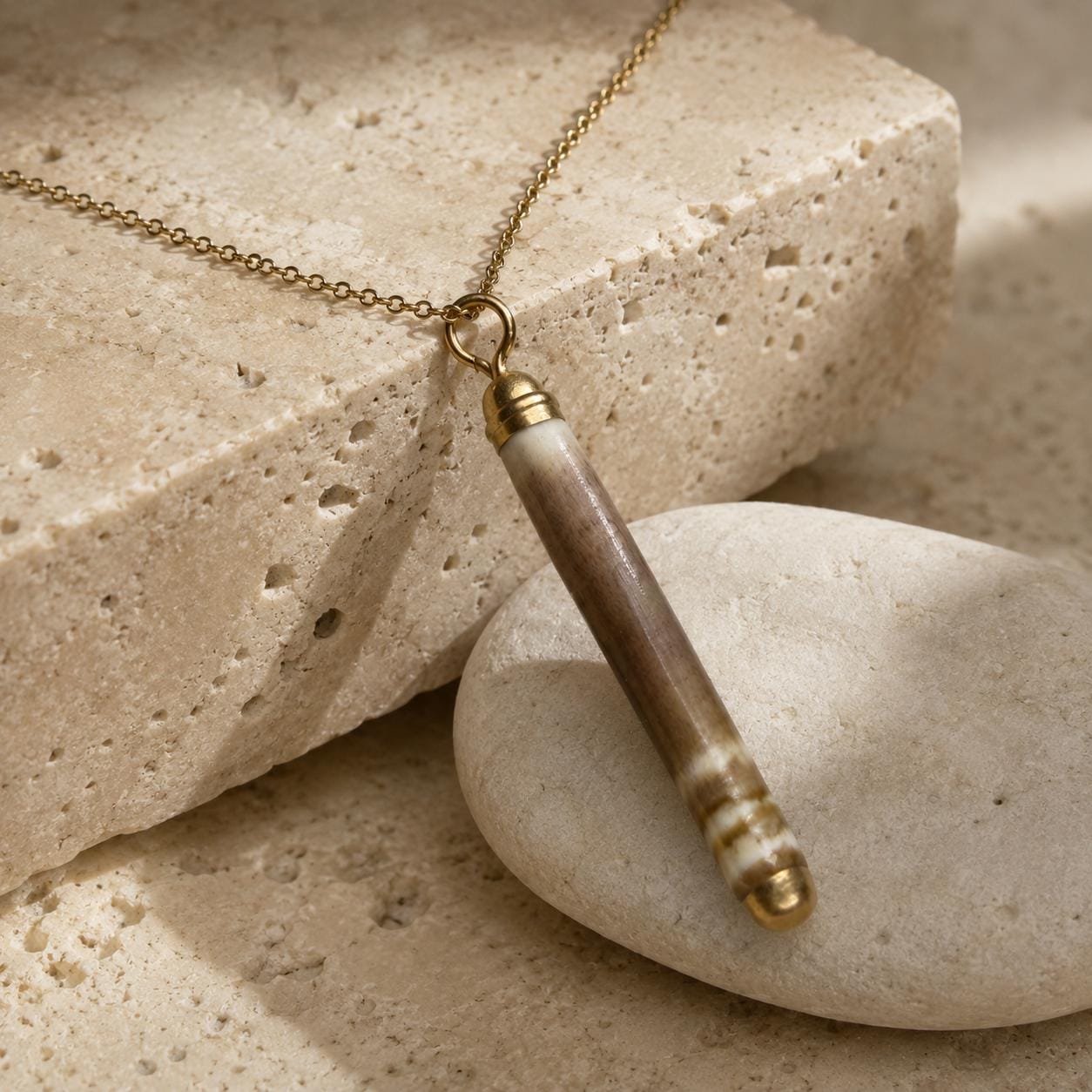 Close-up of a naturally banded sea-urchin spine pendant on beige travertine, showing its organic surface, gold-leaf tips and small attachment rin