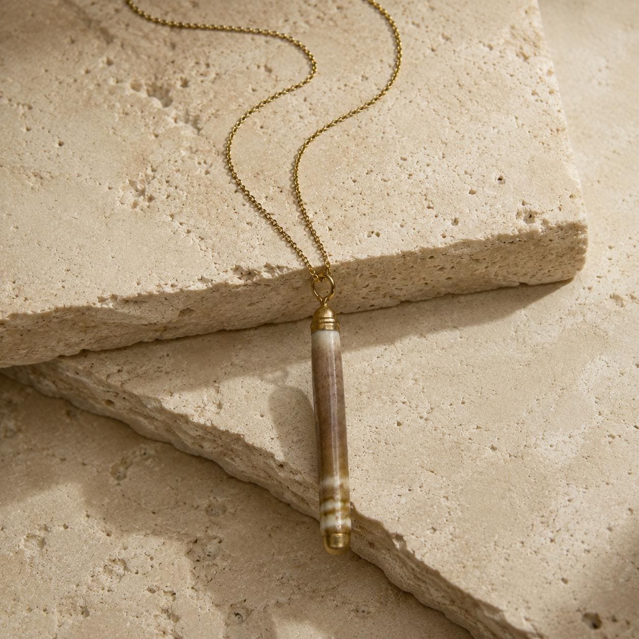 Close-up of a naturally banded sea-urchin spine pendant on beige travertine, showing its organic surface, gold-leaf tips and small attachment rin