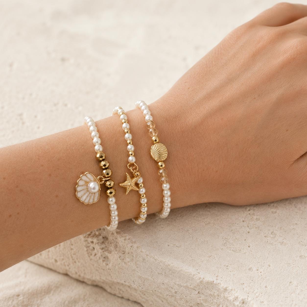 Set of three delicate coastal stretch bracelets featuring small ivory faux pearls and gold-tone seaside details. The coordinating jewellery set includes a blush enamel-style scallop shell charm with pearl centre, a textured gold starfish charm and a ridged gold shell centrepiece with pale champagne faceted beads. Lightweight 16 cm elasticated bohemian costume jewellery for coastal gifts, Christmas presents, stocking fillers, holidays, beach weddings, bridesmaids and gifts for her.