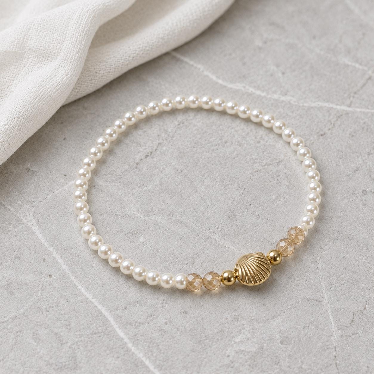 Set of three delicate coastal stretch bracelets featuring small ivory faux pearls and gold-tone seaside details. The coordinating jewellery set includes a blush enamel-style scallop shell charm with pearl centre, a textured gold starfish charm and a ridged gold shell centrepiece with pale champagne faceted beads. Lightweight 16 cm elasticated bohemian costume jewellery for coastal gifts, Christmas presents, stocking fillers, holidays, beach weddings, bridesmaids and gifts for her.