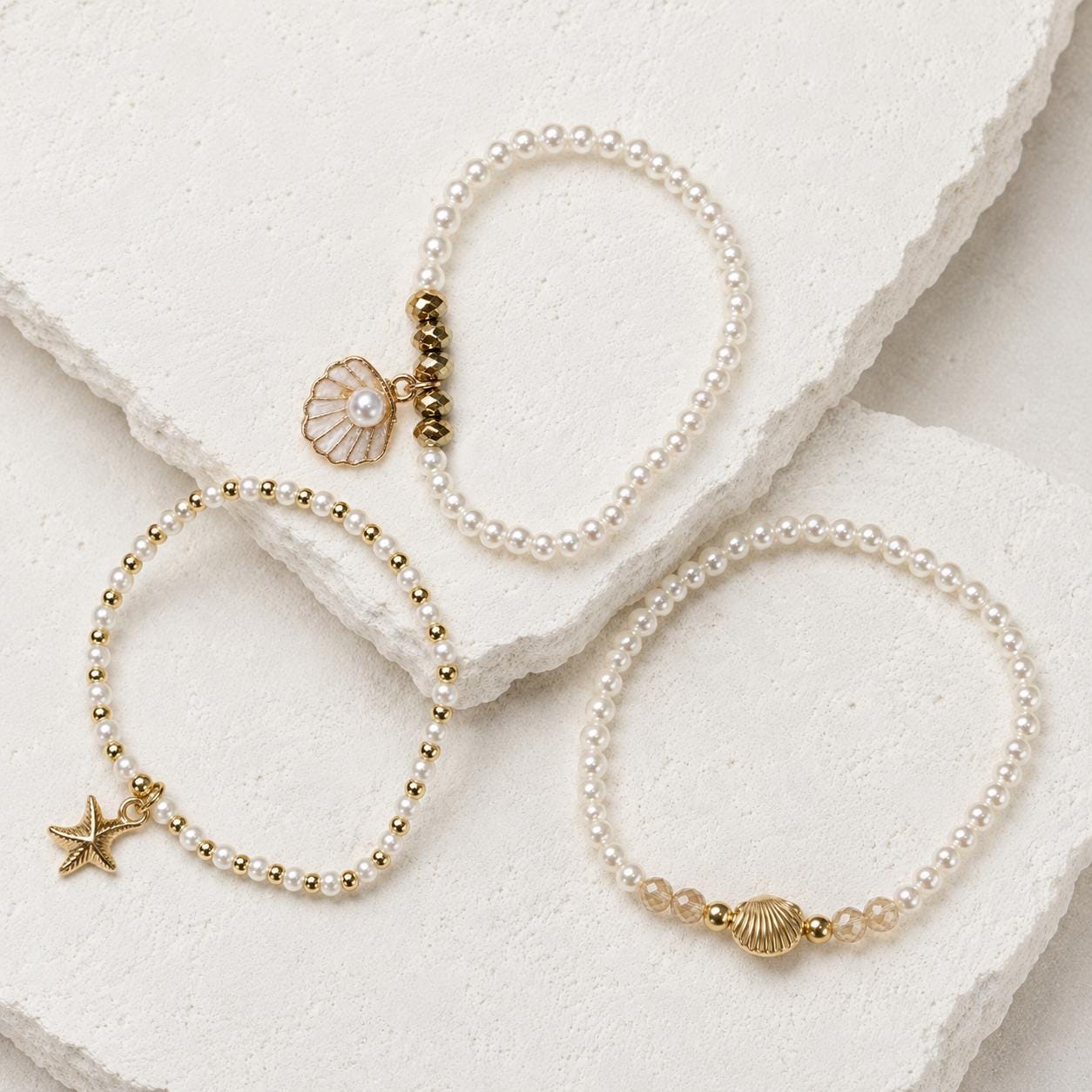 Set of three delicate coastal stretch bracelets featuring small ivory faux pearls and gold-tone seaside details. The coordinating jewellery set includes a blush enamel-style scallop shell charm with pearl centre, a textured gold starfish charm and a ridged gold shell centrepiece with pale champagne faceted beads. Lightweight 16 cm elasticated bohemian costume jewellery for coastal gifts, Christmas presents, stocking fillers, holidays, beach weddings, bridesmaids and gifts for her.