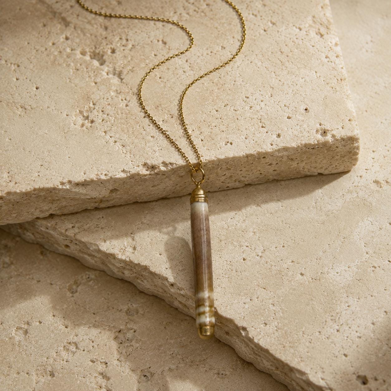 Close-up of a naturally banded sea-urchin spine pendant on beige travertine, showing its organic surface, gold-leaf tips and small attachment rin