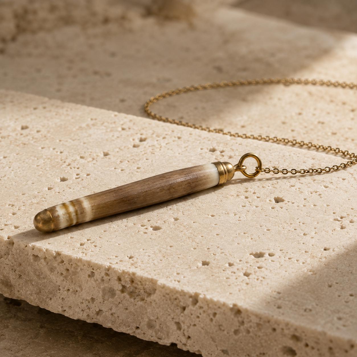 Close-up of a naturally banded sea-urchin spine pendant on beige travertine, showing its organic surface, gold-leaf tips and small attachment rin